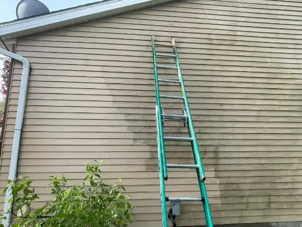 Dirty house siding part way through a wash, with a ladder set against the wall