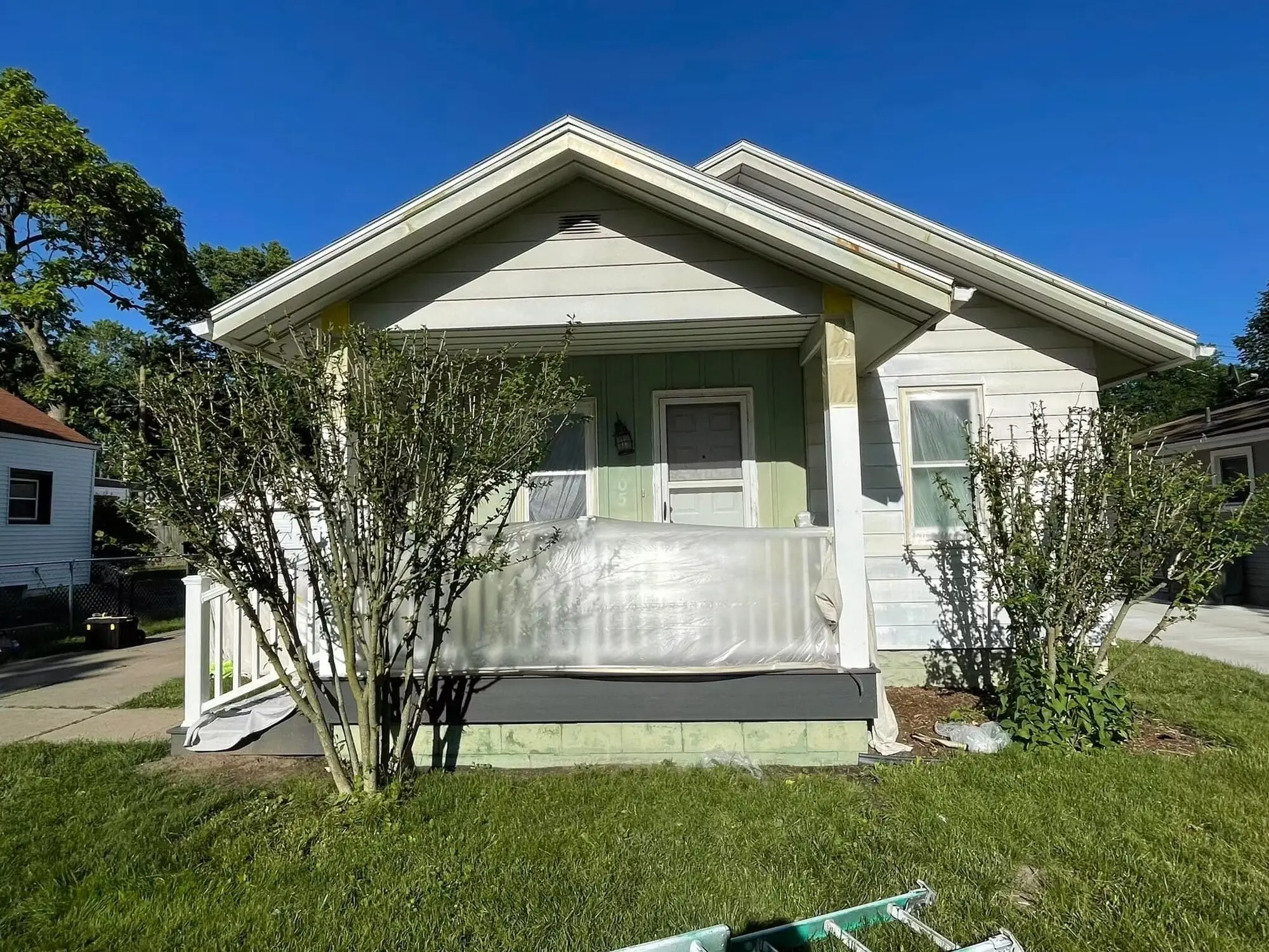 Bungalow with worn green and cream siding, the porch rail wrapped in plastic sheeting before painting