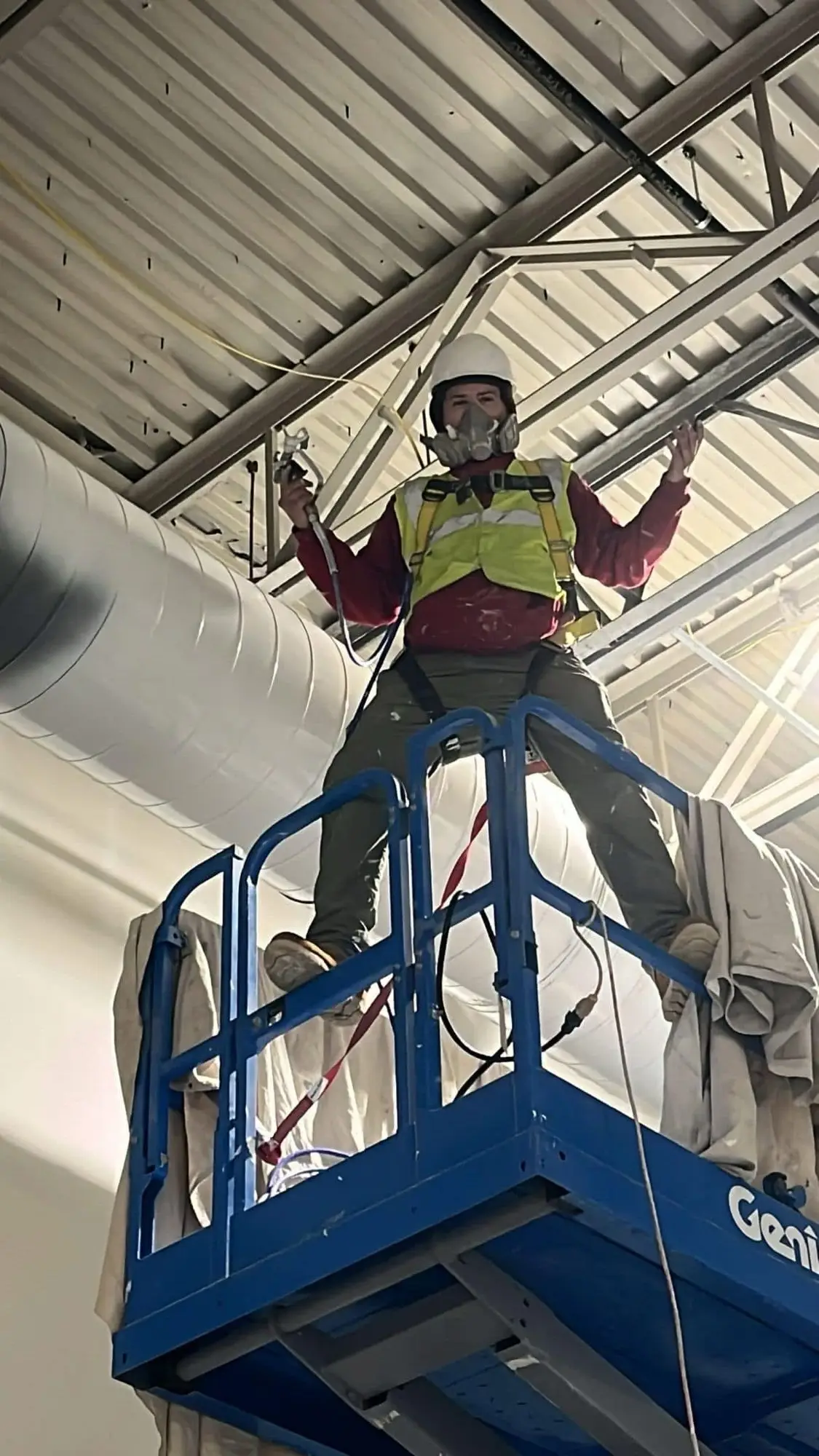 Painter in a hard hat, harness and respirator spraying a warehouse ceiling deck from a scissor lift
