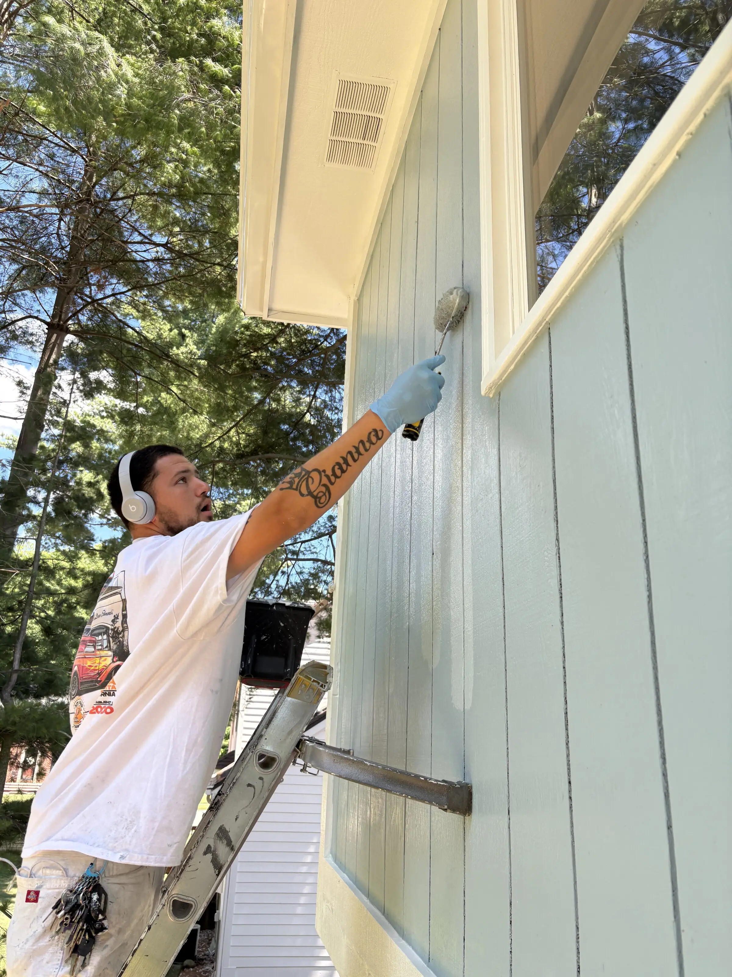 Painter on a ladder rolling pale blue siding up to the cream soffit and window trim